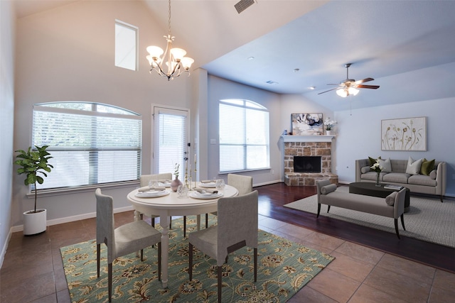 dining area featuring ceiling fan with notable chandelier, a stone fireplace, dark tile patterned floors, and vaulted ceiling