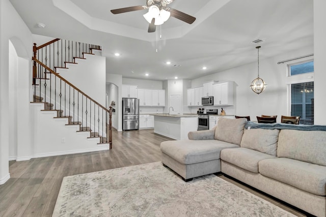 living room featuring a tray ceiling, sink, ceiling fan with notable chandelier, and light wood-type flooring