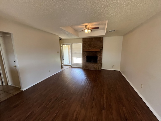 unfurnished living room featuring ceiling fan, a raised ceiling, a brick fireplace, a textured ceiling, and dark wood-type flooring