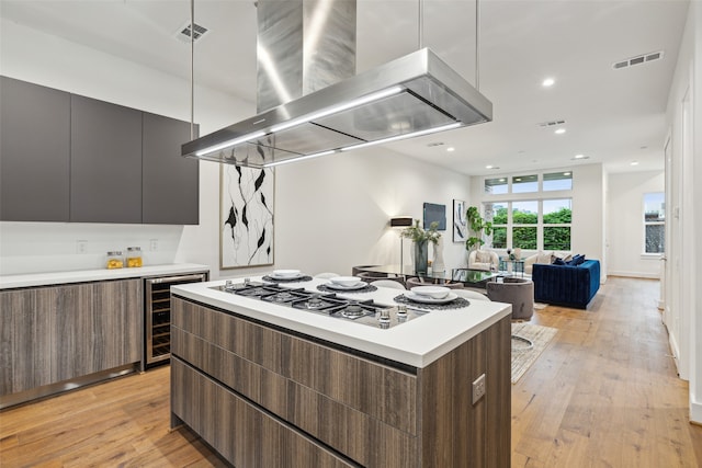 kitchen with dark brown cabinets, beverage cooler, a kitchen island, wall chimney exhaust hood, and light hardwood / wood-style flooring