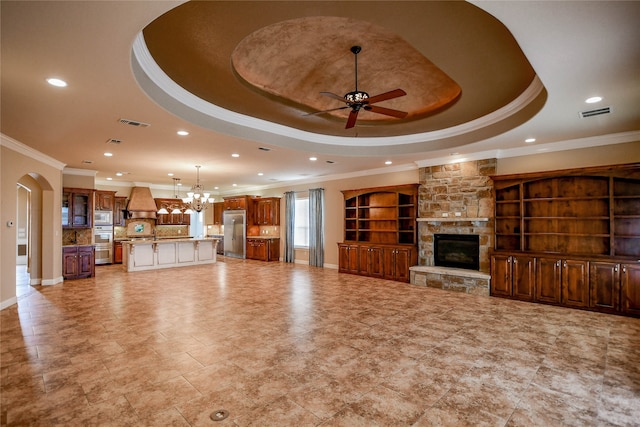 unfurnished living room featuring built in shelves, a raised ceiling, a stone fireplace, ceiling fan with notable chandelier, and crown molding