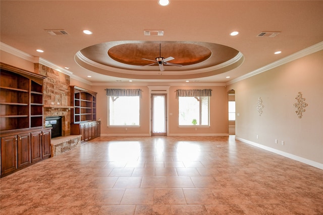 unfurnished living room with a stone fireplace, crown molding, a tray ceiling, ceiling fan, and built in shelves