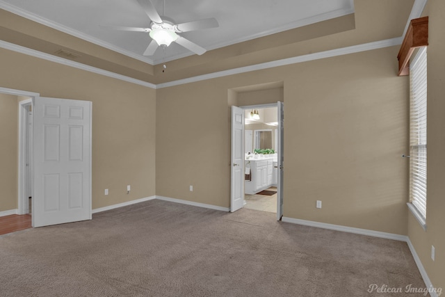 unfurnished bedroom with ceiling fan, a tray ceiling, ensuite bathroom, crown molding, and light colored carpet