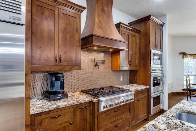 kitchen featuring light stone counters, custom exhaust hood, appliances with stainless steel finishes, and backsplash