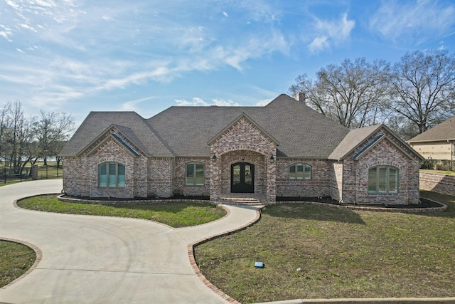 french country home featuring a front lawn, curved driveway, brick siding, and roof with shingles