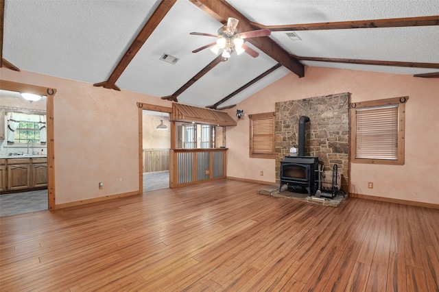 unfurnished living room featuring a wood stove, light wood-type flooring, a textured ceiling, ceiling fan, and vaulted ceiling with beams