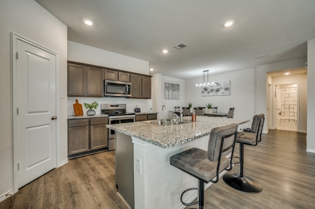 kitchen featuring appliances with stainless steel finishes, an island with sink, light stone countertops, dark hardwood / wood-style flooring, and a kitchen bar