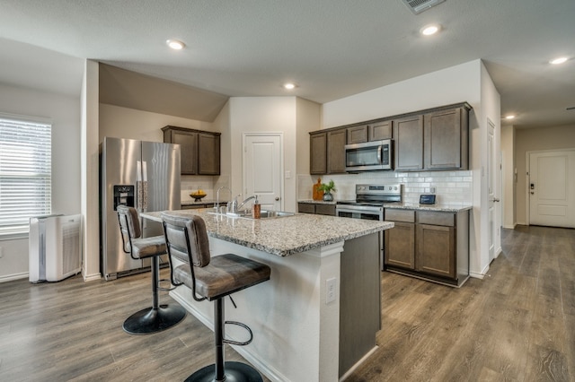 kitchen with dark wood-type flooring, a kitchen island with sink, sink, appliances with stainless steel finishes, and light stone countertops