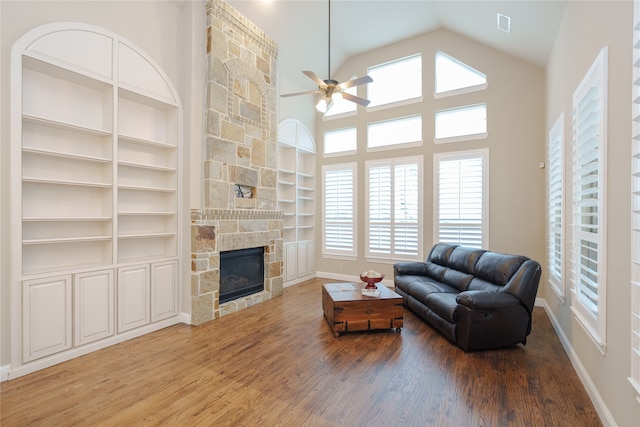 living room with built in shelves, ceiling fan, wood-type flooring, a stone fireplace, and high vaulted ceiling