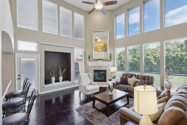 living room with a high ceiling, a tiled fireplace, ceiling fan, and dark wood-type flooring