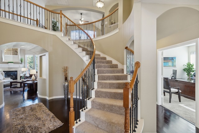 staircase featuring a towering ceiling, ceiling fan, a tile fireplace, and hardwood / wood-style floors