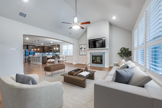 living area with visible vents, a tile fireplace, light wood-style flooring, high vaulted ceiling, and ceiling fan with notable chandelier