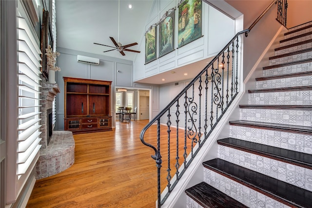 staircase featuring hardwood / wood-style floors, a wall mounted AC, ceiling fan, high vaulted ceiling, and a fireplace