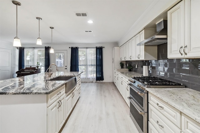 kitchen featuring light stone counters, wall chimney exhaust hood, sink, a center island with sink, and range with two ovens