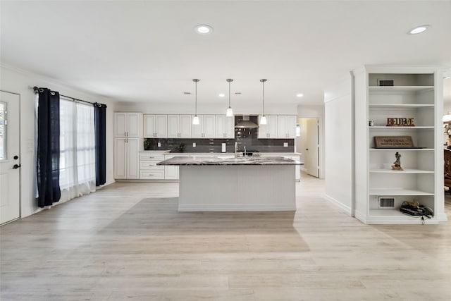 kitchen featuring pendant lighting, wall chimney range hood, light hardwood / wood-style flooring, an island with sink, and white cabinetry