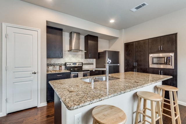 kitchen with sink, wall chimney range hood, a center island with sink, appliances with stainless steel finishes, and dark hardwood / wood-style flooring