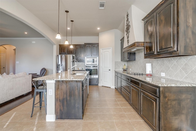 kitchen with hanging light fixtures, an island with sink, backsplash, stainless steel appliances, and dark brown cabinets