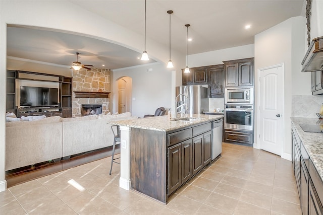 kitchen featuring dark brown cabinets, a center island with sink, appliances with stainless steel finishes, a stone fireplace, and ceiling fan