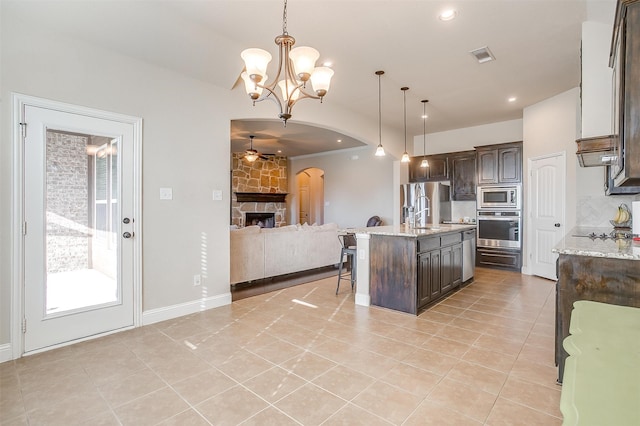 kitchen with an island with sink, dark brown cabinets, stainless steel appliances, a fireplace, and ceiling fan with notable chandelier