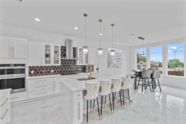 kitchen with wall chimney exhaust hood, stainless steel double oven, a kitchen island with sink, sink, and white cabinets