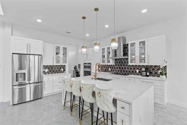 kitchen with wall chimney exhaust hood, backsplash, a kitchen island with sink, white cabinets, and appliances with stainless steel finishes