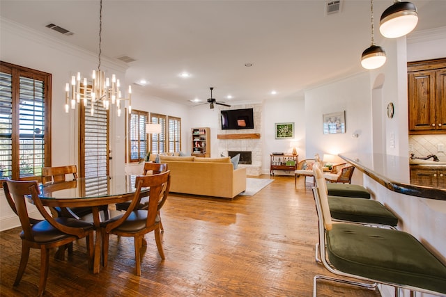 dining area with ceiling fan with notable chandelier, light wood-type flooring, crown molding, and a stone fireplace