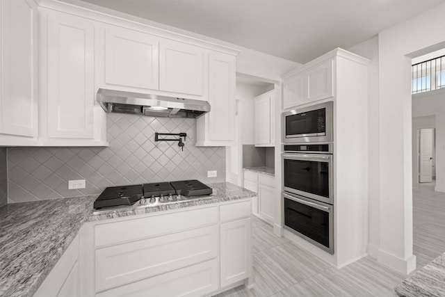 kitchen featuring white cabinetry, backsplash, range hood, stainless steel appliances, and light stone countertops