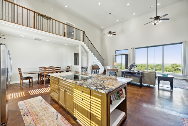 kitchen with black electric stovetop, a high ceiling, a center island, ceiling fan, and dark stone counters