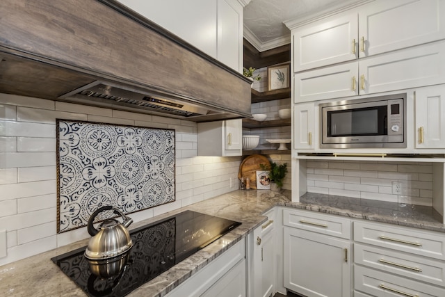 kitchen with light stone countertops, decorative backsplash, stainless steel microwave, white cabinetry, and black electric stovetop