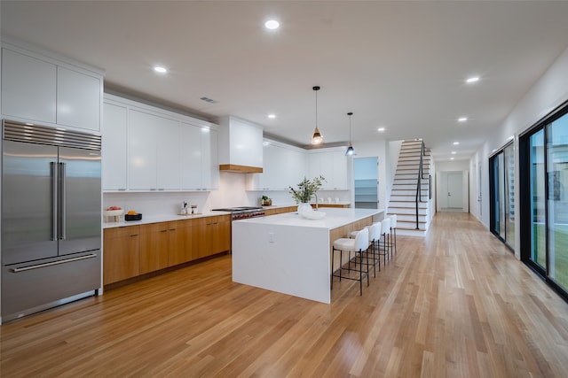 kitchen with stainless steel built in fridge, a kitchen island, white cabinetry, light wood-type flooring, and decorative light fixtures