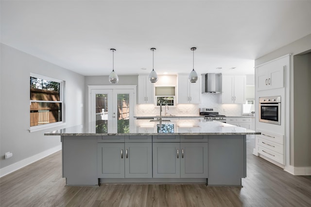 kitchen with light stone counters, a center island with sink, wall chimney exhaust hood, white cabinetry, and appliances with stainless steel finishes