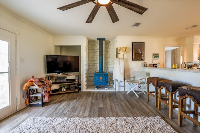 living room with wood-type flooring, sink, a wood stove, crown molding, and ceiling fan