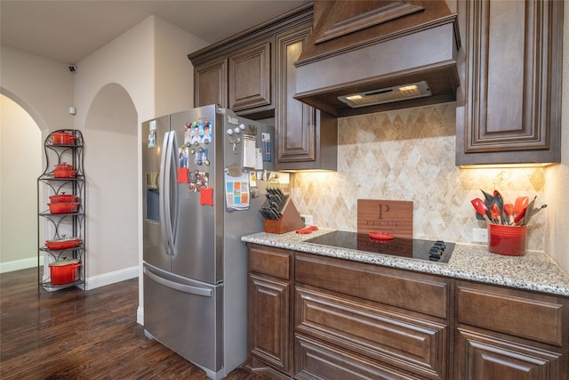 kitchen featuring light stone counters, stainless steel fridge, custom range hood, black electric stovetop, and decorative backsplash