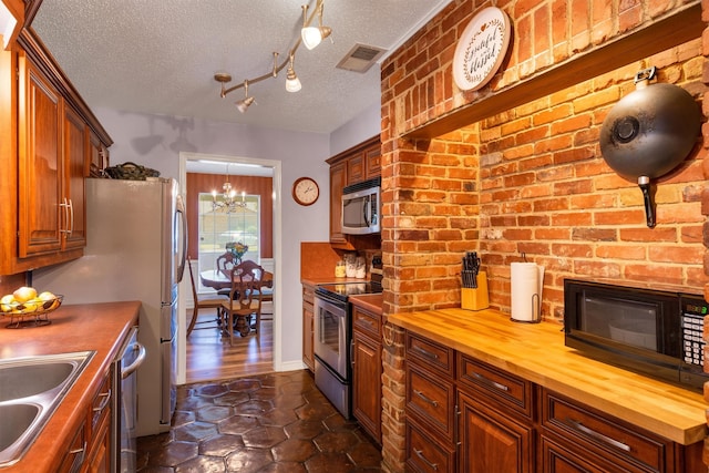 kitchen with brick wall, butcher block countertops, stainless steel appliances, a textured ceiling, and an inviting chandelier