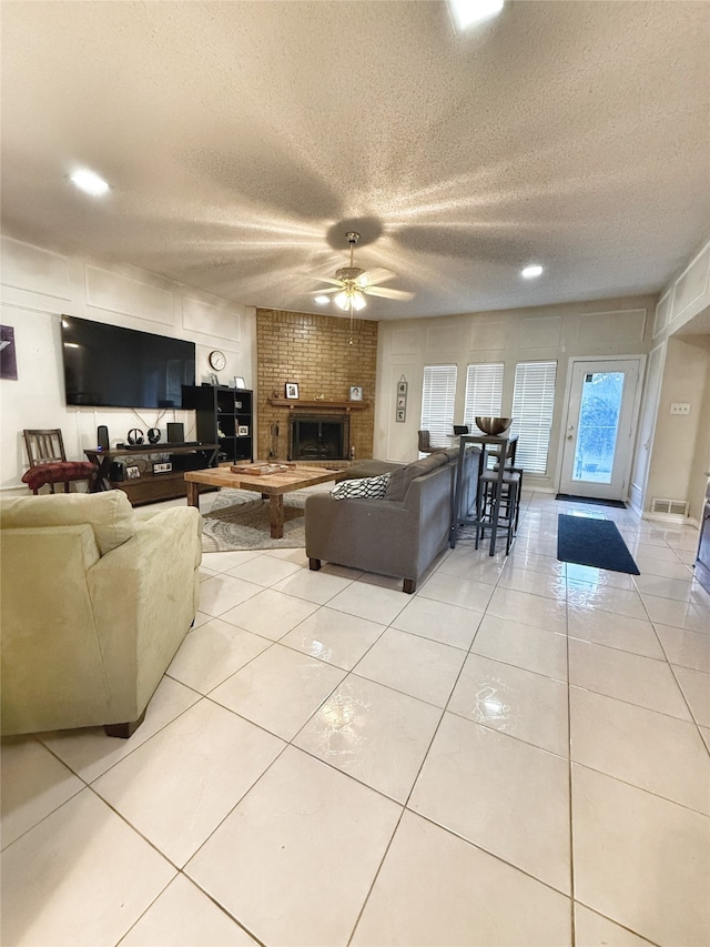 living room with ceiling fan, a textured ceiling, a brick fireplace, and light tile patterned flooring