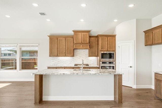 kitchen with light stone counters, wood-type flooring, stainless steel appliances, and an island with sink