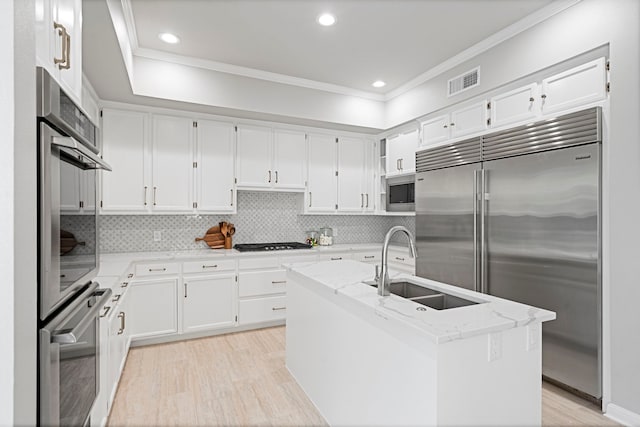 kitchen featuring light stone counters, a kitchen island with sink, built in appliances, sink, and white cabinets