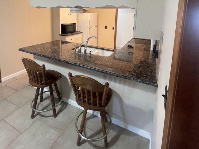 kitchen featuring black microwave, a peninsula, a sink, freestanding refrigerator, and dark stone counters