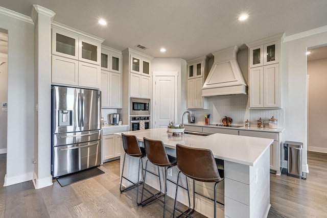 kitchen with custom exhaust hood, an island with sink, stainless steel appliances, and ornamental molding