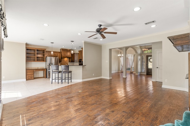 unfurnished living room featuring built in desk, crown molding, ceiling fan with notable chandelier, and light wood-type flooring