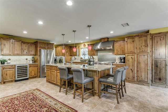 kitchen featuring wall chimney range hood, stainless steel fridge, a kitchen island with sink, washing machine and dryer, and beverage cooler