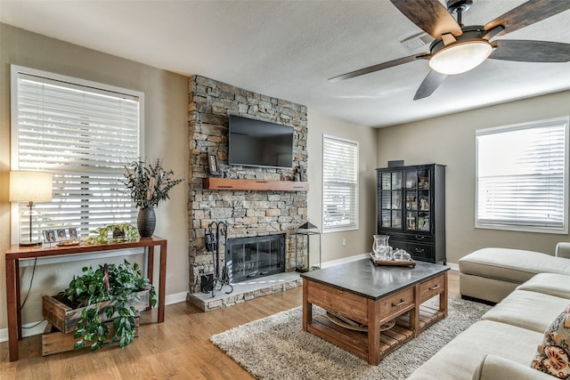 living room featuring ceiling fan, a stone fireplace, a textured ceiling, and light hardwood / wood-style floors