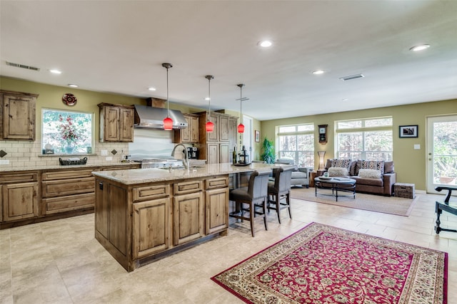 kitchen featuring pendant lighting, a kitchen breakfast bar, decorative backsplash, a kitchen island with sink, and wall chimney exhaust hood