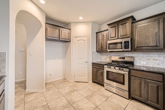 kitchen featuring tasteful backsplash, stainless steel appliances, light tile patterned floors, dark brown cabinetry, and dark stone countertops