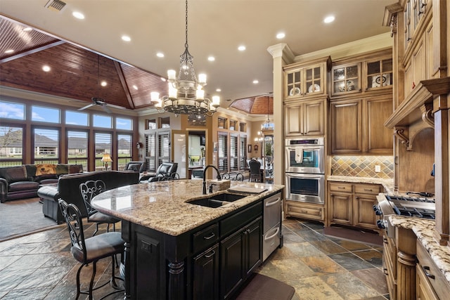 kitchen with sink, ceiling fan with notable chandelier, an island with sink, stainless steel appliances, and light stone countertops