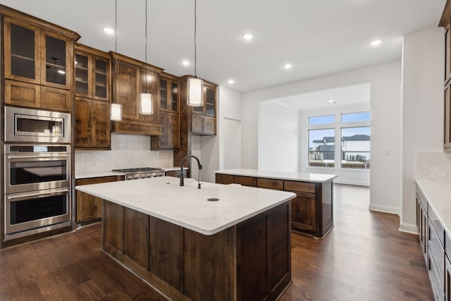 kitchen featuring decorative backsplash, appliances with stainless steel finishes, light stone counters, a kitchen island with sink, and pendant lighting