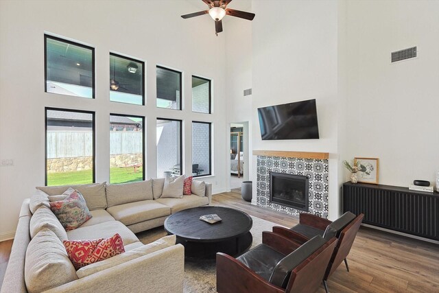 living room with ceiling fan, a tiled fireplace, a high ceiling, and hardwood / wood-style flooring