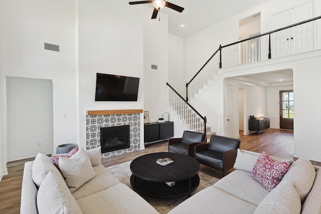 living room featuring a tiled fireplace, ceiling fan, wood-type flooring, and a towering ceiling