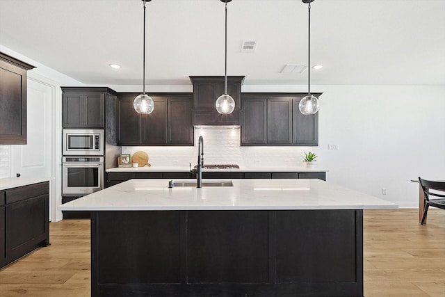 kitchen featuring appliances with stainless steel finishes, dark brown cabinetry, hanging light fixtures, a kitchen island with sink, and light wood-type flooring