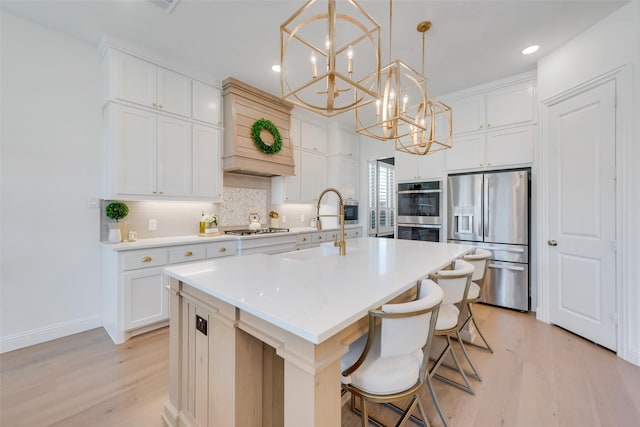 kitchen featuring appliances with stainless steel finishes, light wood-type flooring, white cabinetry, hanging light fixtures, and an island with sink